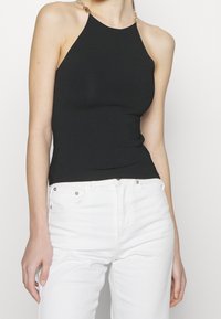 Schwarzes Neckholder-Oberteil aus geripptem Material mit höherem Ausschnitt und goldenen Kettenakzenten an den Schultern, kombiniert mit weißen Jeans.