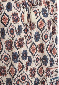 Luchtige blouse met een beige achtergrond, versierd met bloem- en geometrische patronen in rood en marineblauw, afgewerkt met witte franje bij de hals.