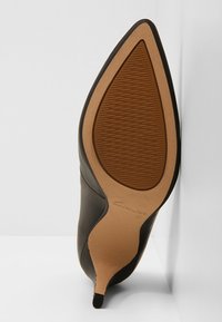 Chaussure à talon aiguille noire à bout pointu avec semelle texturée couleur tan et petite pointe de talon, inclinée contre un mur blanc d'angle.