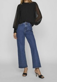 Top trasparente nero con maniche a palloncino abbinato a jeans blu a vita alta con gamba larga e sandali neri con tacco a blocco.