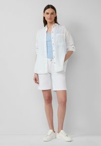 Witte linnen overhemd met knopen over een blauwe tanktop, gecombineerd met witte opgerolde shorts en witte sneakers. Eenvoudige, casual outfit.