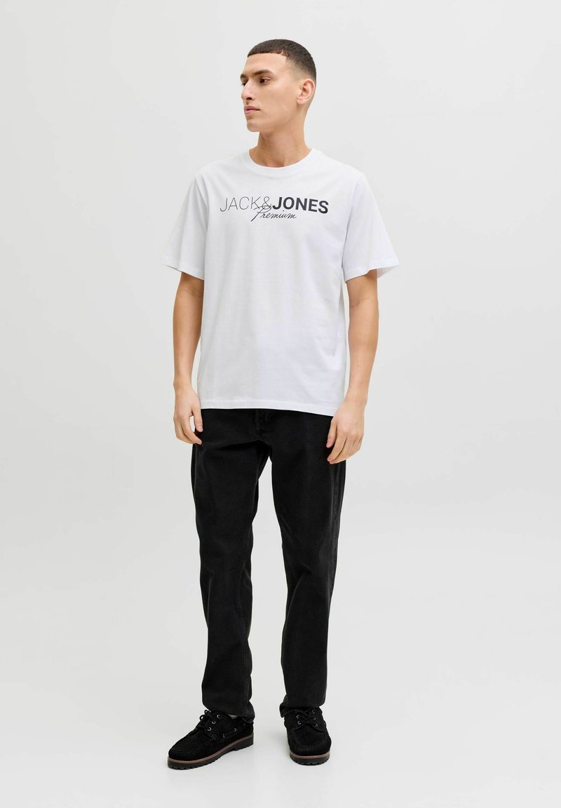 Young man standing wearing white "Jack & Jones Premium" T-shirt, black pants, and black shoes, facing slightly left on plain background.