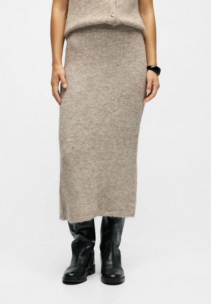 Jupe en mélange de laine tricotée de couleur marron clair avec une forme droite et une taille côtelée. Associée à des bottes en cuir noires.