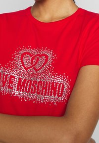 Rød t-shirt med et dekorativt design af rhinsten, der danner hjerter og ordet "Moschino", med en tætsiddende og blød tekstur.