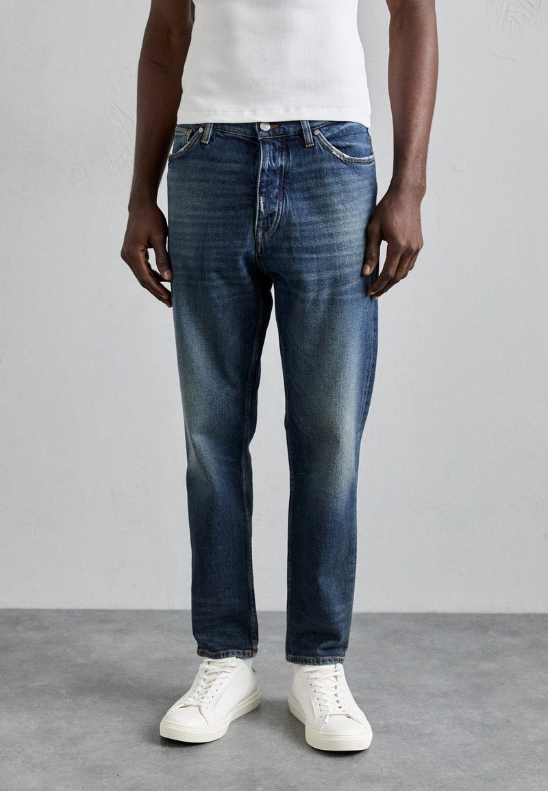 Homme portant un jean bleu délavé, des baskets blanches et un débardeur blanc, debout sur un sol gris contre un mur uni.