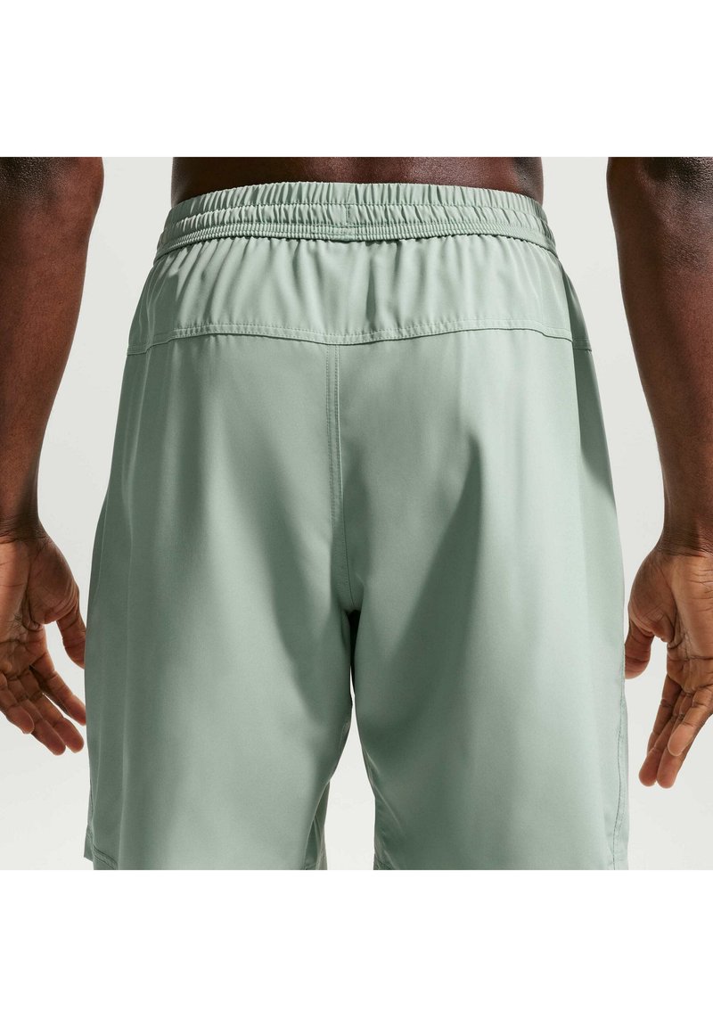 Gros plan d’une personne portant un short de sport vert clair avec une taille élastique, vue de dos de la taille jusqu’au milieu de la cuisse, les mains détendues le long du corps.