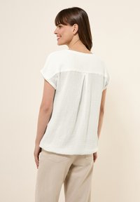 Hvid kortærmet top lavet af tekstureret stof med en løs pasform, der har en detalje med en plissering bagpå. Kombinationen med lyse beige bukser.
