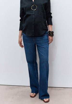 Dark blue flared jeans made of denim, paired with a black button-up shirt, a large gold ring necklace, and black bangles.