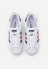 Scarpe sportive bianche con una punta testurizzata, caratterizzate da strisce nere, rosse e blu navy. Lacci e logo del marchio sulla linguetta.