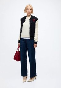 Femme aux cheveux blonds courts, debout, portant une veste universitaire noire et blanche, un haut blanc, un jean foncé, des talons beiges et tenant un sac à main rouge.