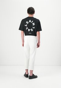 Czarny bawełniany t-shirt z dużym białym okręgiem i tekstem z tyłu; zestawiony z białymi, skróconymi spodniami i czarnymi trampkami. Prosty design, casualowy styl.
