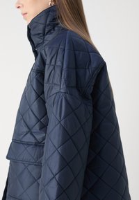 Ναυτικό επενδεδυμένο μπουφάν με γούνα που διαθέτει όρθιο γιακά, διαμαντένια ραφή και φαρδιά μανίκια, κατασκευασμένο από απαλή, γυαλιστερή ύφασμα.