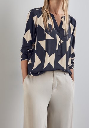 Woman wearing a black and beige geometric patterned blouse with long sleeves and beige high-waist pants, hands in pockets, neutral background.