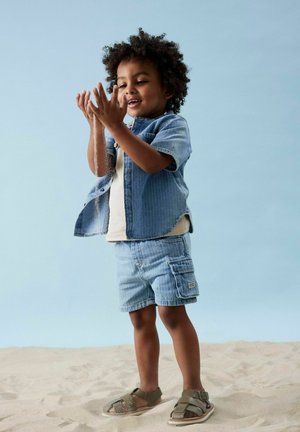 Denim shirt en cargo shorts in lichtblauw, gecombineerd met grijze sandalen. Kind speelt met zand tegen een lichtblauwe achtergrond.
