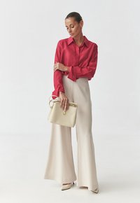 Chemise rouge en satin à boutons, pantalon large crème, et sac à main crème avec quincaillerie dorée. Des talons beiges pointus achèvent le look.