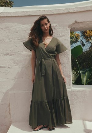 Femeie cu păr lung și ondulat, purtând o rochie tip halat verde măsliniu și un colier din scoici, stând în fața unui perete texturat alb, cu o fereastră care arată plante.