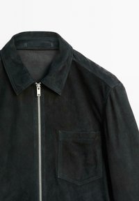 Schwarze Wildlederjacke mit Kragen, Reißverschluss vorne und einer Brusttasche. Glatte Textur mit mattem Finish; Schulterakzente aus passendem Wildleder.
