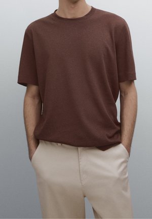 Personne portant une chemise marron unie à manches courtes rentrée dans un pantalon beige clair, les mains dans les poches, sur fond gris.
