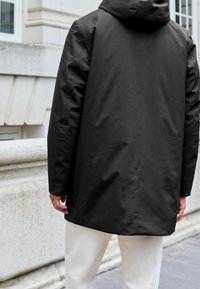 Czarny, wodoodporny płaszcz z kapturem; ma prosty krój i gładką fakturę. Widok od tyłu podkreśla detale szwów i minimalistyczny design.