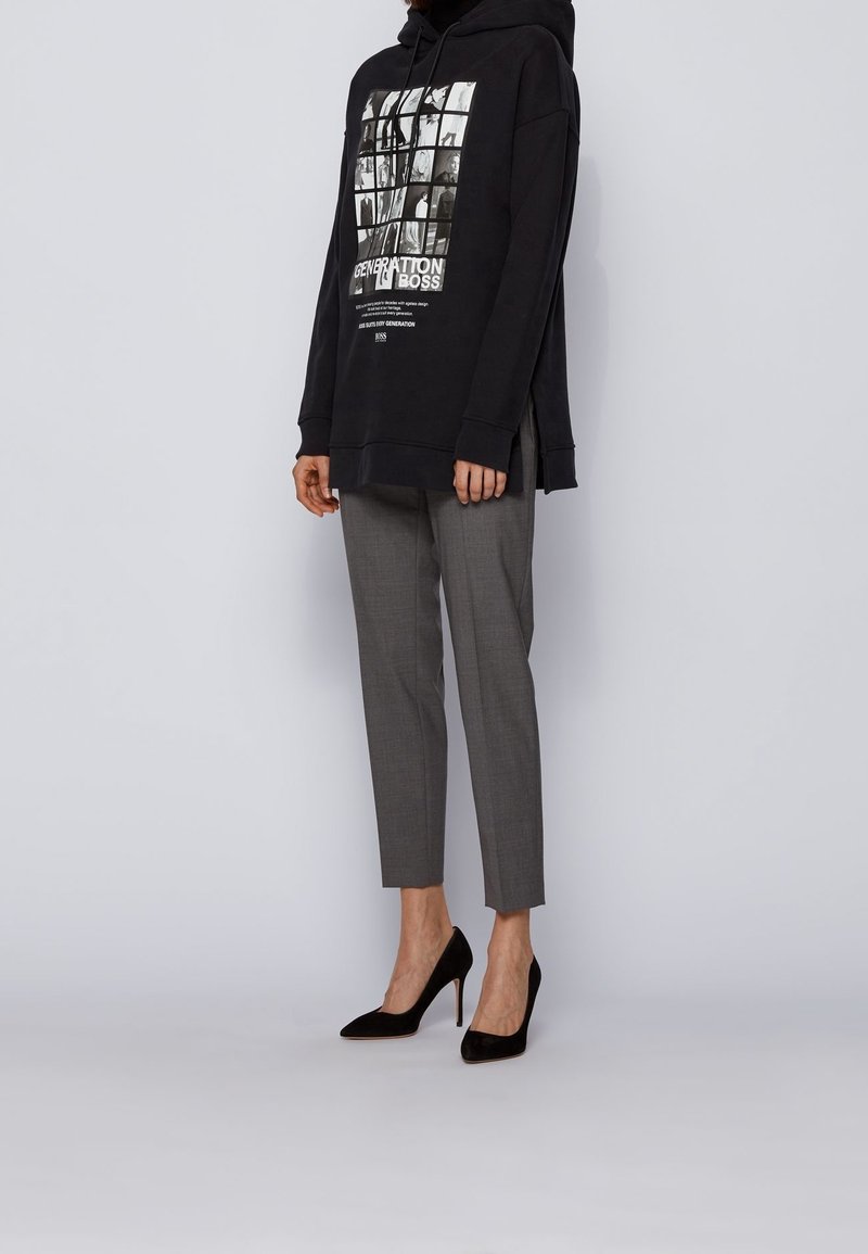 Černá mikina s kapucí a grafickým potiskem, kombinovaná se šedými přizpůsobenými kalhotami a černými lodičkami na vysokém podpatku. Hladká tkanina a elegantní design.