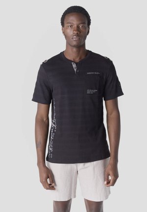 Férfi fekete rövid ujjú Henley pólóval, rajta szöveggel, bézs rövidnadrágban, világosszürke sima háttér előtt áll.