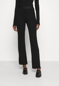 Czarne ryflowane spodnie dzianinowe z rozszerzanymi nogawkami i postrzępionymi brzegami, z dopasowanym pasem w talii. Materiał ma gładką strukturę. Połączone z czarnymi butami.