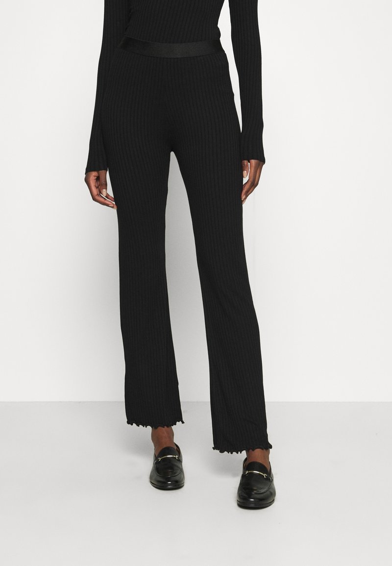 Czarne ryflowane spodnie dzianinowe z rozszerzanymi nogawkami i postrzępionymi brzegami, z dopasowanym pasem w talii. Materiał ma gładką strukturę. Połączone z czarnymi butami.