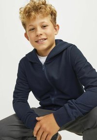 Felpa con zip di colore blu navy in morbido tessuto, dotata di cappuccio, polsini a costine e vestibilità casual, abbinata a pantaloni grigi.