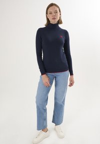 Maglione a collo alto blu navy con un logo a forma di cuore rosso sul petto, abbinato a jeans dritti azzurri chiari e sneakers bianche. Materiale in maglia elasticizzata.
