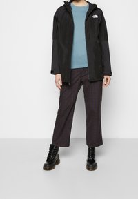 Czarna wodoodporna kurtka z kapturem, pod spodem niebieska koszulka, w kratę brązowe spodnie. Strój dopełniają czarne sznurowane buty.