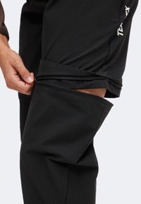 Main déboutonnant un pantalon convertible noir, montrant la partie inférieure détachée de la partie supérieure, avec une jambe partiellement visible à l'intérieur.