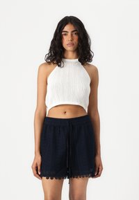 Hvitt strikket crop top med høy hals kombinert med mørkeblå shorts med intrikate mønstre og en blondekant nederst.