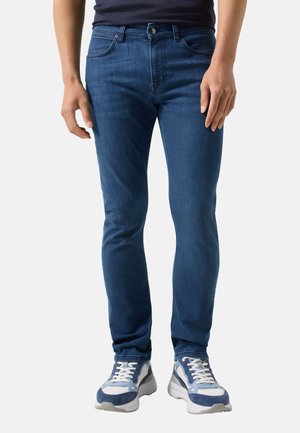 Mężczyzna w obcisłych niebieskich dżinsach i białych z niebieskimi elementami sportowych butach, stojący na tle jasnego, jednolitego tła.