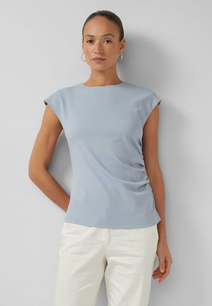 Vrouw die een lichtblauwe mouwloze top en witte broek draagt, staat tegen een grijze muur met één hand achter haar rug.