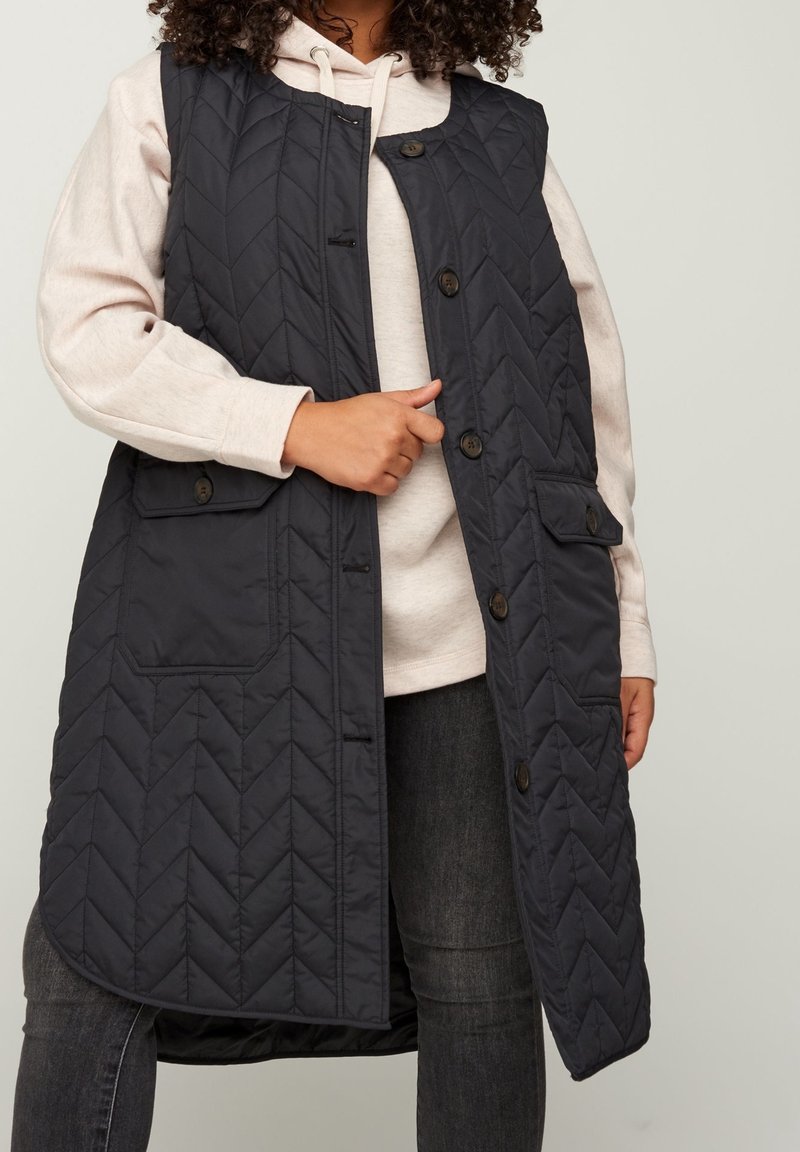 Gilet matelassé sans manches en bleu marine foncé, avec un motif en chevron, des boutons de fermeture et deux poches avant, porté par-dessus un hoodie clair.