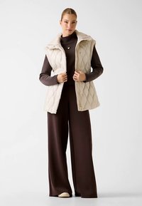Gilet trapuntato beige con colletto a listino e bottoni a scatto, indossato sopra una maglia a manica lunga marrone scuro e pantaloni a gamba larga. Tessuto dalla texture liscia.