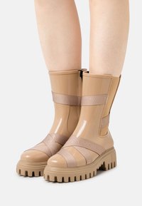 Botins bejes até ao tornozelo apresentam um acabamento brilhante, lados elásticos e solas de borracha largas com ranhuras texturizadas. Correias cruzadas exibem detalhes do logotipo.