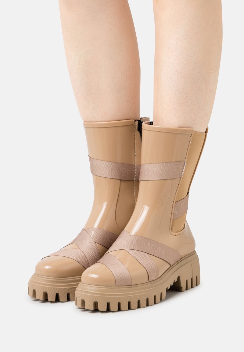 Botins bejes até ao tornozelo apresentam um acabamento brilhante, lados elásticos e solas de borracha largas com ranhuras texturizadas. Correias cruzadas exibem detalhes do logotipo.