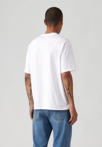 Balta medvilninė t-shirt su trumpomis rankovėmis ir tiesiu kirpimu, derinama su šviesiai mėlynais džinsais. Matomi tatuiruotės ant rankos.