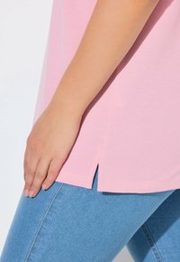 Jasnoróżowa bluzka z krótkim rękawem z bocznymi rozcięciami, teksturowanym materiałem i luźnym krojem, zestawiona z niebieskimi dżinsami.