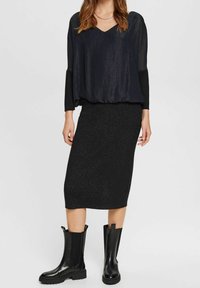 Top lungo a maniche lunghe nero e strutturato con scollatura a V, abbinato a una gonna midi nera aderente e luccicante. Indossato con stivali neri fino al ginocchio.