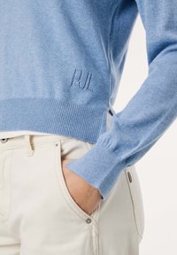 Svetlo modri pleteni pulover z rebrastim robom in manšetami, ki ima na sprednji strani subtilno vzorčast "PUL" logotip; oblečen z smetanovimi hlačami.