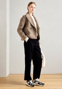 Vrouw die binnen staat en een beige cropped dubbelzijdige jas, een roomkleurige trui, zwarte wijde broek, zwart-witte sneakers en een witte tas draagt.