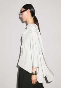 Blouse drapée de couleur claire avec un col haut, des manches longues et des détails plissés. Le mannequin porte des bracelets en métal argenté et des lunettes.