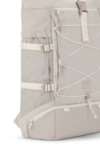 Beige hátizsák texturált anyagból, több cipzáras rekesszel, oldalsó hevederekkel és rugalmas bungee zsinórral. Kiemelt logóval ellátva.