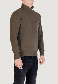 Uomo che indossa un maglione a collo alto lavorato a maglia marrone e jeans neri, in piedi contro uno sfondo chiaro e uniforme.