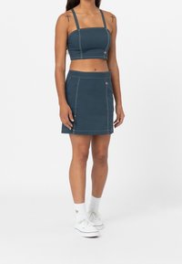 Tmavě tyrkysový dvoudílný outfit skládající se z krátkého topu s tenkými ramínky a odpovídající mini sukně, která má kontrastní švy.