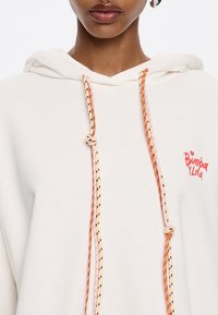 Sudadera crema con un bolsillo frontal, cordones ajustables en naranja y rosa, y el logo de "Bimba Y Lola" en rojo en el lado izquierdo.