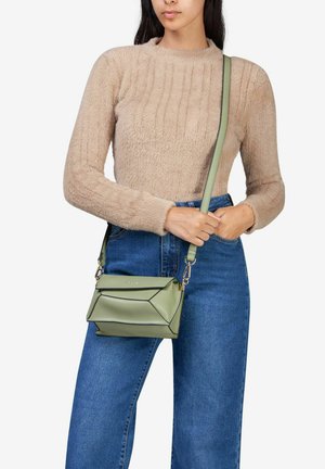 Grøn læder crossover taske med geometrisk design, sort kantbånd og justerbar rem, holdt foran en person iført en beige sweater.