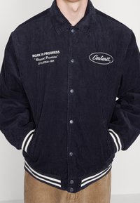 Giacca in coste di velluto blu navy con bottoni a pressione, loghi ricamati bianchi e polsini e orlo a righe. Presenta una vestibilità rilassata e una texture evidente.
